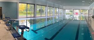 Ein Hallenbad mit klarem blauem Wasser und Glaswänden, das die Umgebung draußen reflektiert. Eine Person steht auf einer Leiter auf der rechten Seite.