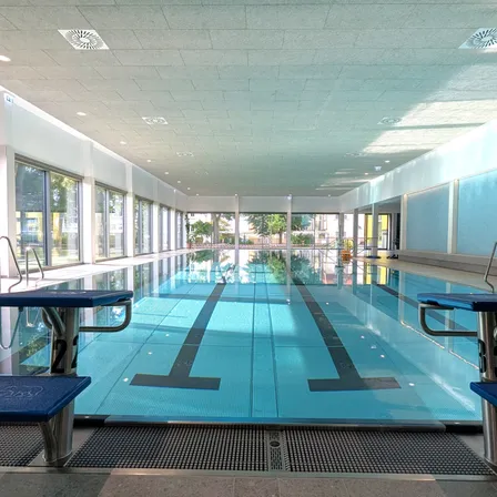 Ein Hallenbad mit klarem blauem Wasser, Bahnmarkierungen und Bänken auf beiden Seiten. Das Becken hat Glaswände und eine Decke mit Beleuchtung. Draußen sind Bäume und Gebäude zu sehen.