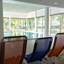 Drei Liegestühle stehen in der Nähe eines Pools, mit großen Fenstern, die auf einen ruhigen Außenbereich blicken. Der Poolbereich hat eine Reflexion von Bäumen, und eine Topfpflanze befindet sich neben dem Fenster.