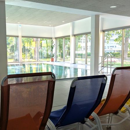 Drei Liegestühle stehen in der Nähe eines Pools, mit großen Fenstern, die auf einen ruhigen Außenbereich blicken. Der Poolbereich hat eine Reflexion von Bäumen, und eine Topfpflanze befindet sich neben dem Fenster.
