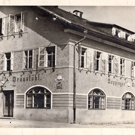 Ein Schwarz-Weiß-Foto aus alten Zeiten zeigt ein Gebäude mit dem Namen Braustube, mit Rundbogenfenstern, einem Schornstein und einem Schild an der Wand.