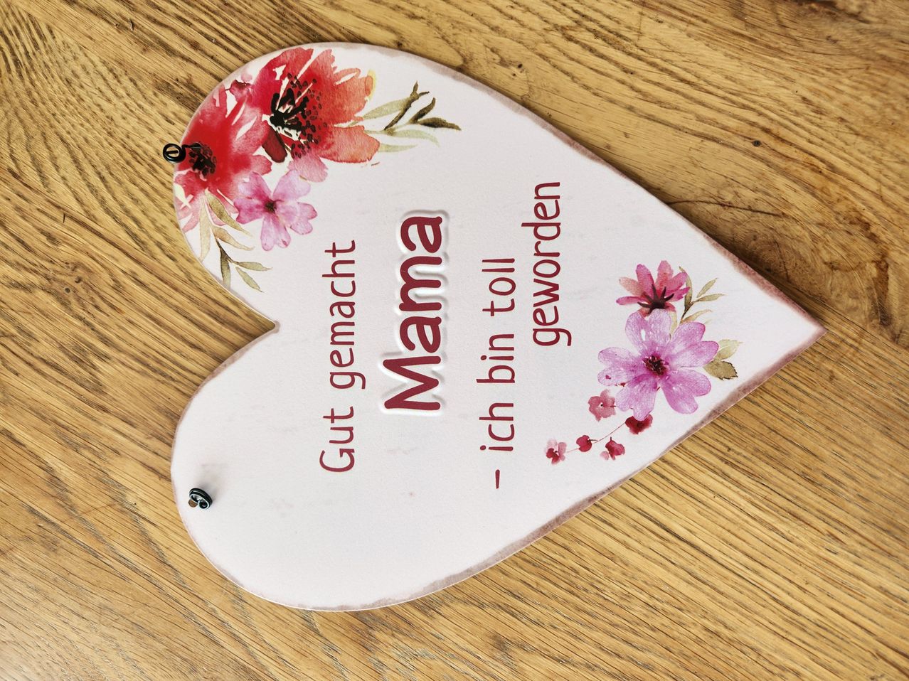 Ein herzförmiges Deko-Stück mit 'Gut gemacht Mama' in Rot geschrieben. Es hat rosa Blumen an den Rändern und steht auf einer Holzoberfläche.