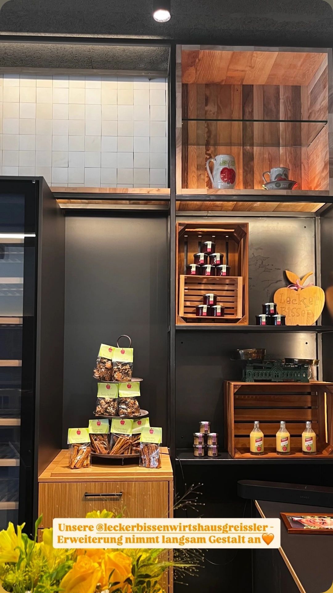Eine Ausstellung verschiedener Lebensmittel in einem Geschäft, einschließlich eines Stapels verpackter Snacks, Gläser mit Konfitüren und Flaschen mit Saft. Holzregale und eine geflieste Wand sind im Hintergrund zu sehen.