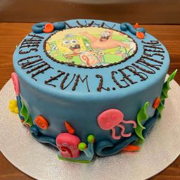 Ein SpongeBob-Kuchen mit blauer Glasur, der ein rundes Bild von SpongeBob und seinen Freunden auf der Oberseite zeigt, umgeben von Meerestieren. Der Kuchen sagt 'Alles Gute zum Geburtstag Luan' in Schokoladenglasur.