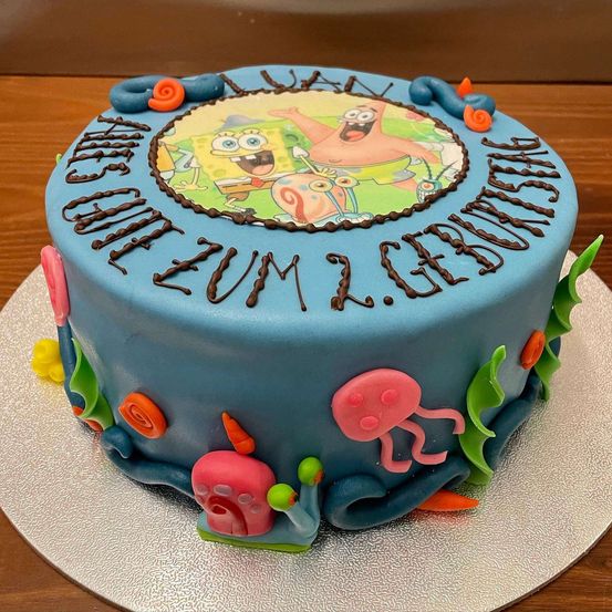 Ein SpongeBob-Kuchen mit blauer Glasur, der ein rundes Bild von SpongeBob und seinen Freunden auf der Oberseite zeigt, umgeben von Meerestieren. Der Kuchen sagt 'Alles Gute zum Geburtstag Luan' in Schokoladenglasur.