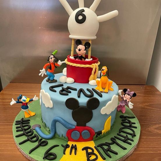 Ein Geburtstagskuchen mit einer 6 auf der Spitze, Mickey Mouse in einem Korb und einem Zug unten. Er hat ein blaues und rotes Thema mit Disney-Charakteren drumherum.