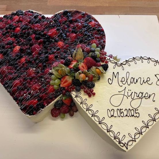 Ein herzförmiger Kuchen mit verschiedenen Beeren und Trauben auf einer weißen Oberfläche. Ein weißer herzförmiger Kuchen mit den Worten Melanie & Jürgen und dem Datum 02.05.2025.