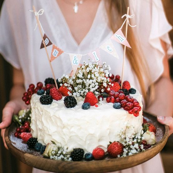 Eine Person hält einen Kuchen mit weißer Glasur und verschiedenen Beeren auf einem Holzteller, verziert mit kleinen Flaggen.