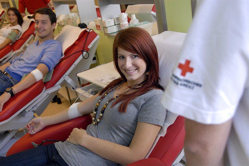 Eine Frau und ein Mann sitzen in roten Stühlen in einem Blutspendezentrum und lächeln.