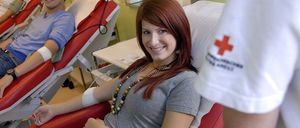 Eine Frau und ein Mann sitzen in roten Stühlen in einem Blutspendezentrum und lächeln.