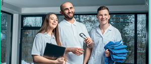 Drei Gesundheitsfachkräfte stehen in einem Raum. Eine Frau hält eine Mappe, und ein Mann neben ihr hält eine Schere. Der andere Mann hält einen Stapel blauer Tücher. Das Bild hat einen Textüberlagerung, der 'Beratungsstelle für Gesundheitsbildung' lautet.