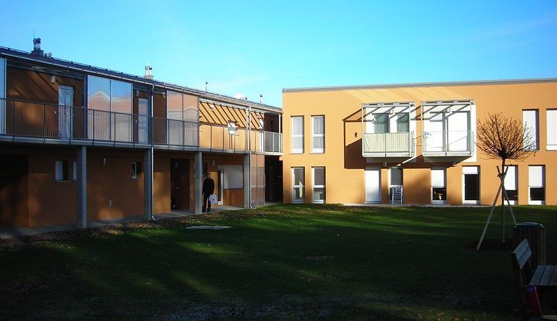Ein gelbes Apartmentgebäude mit Balkonen und einer Person, die davor geht. Ein grüner Rasen und ein kahler Baum sind ebenfalls sichtbar.