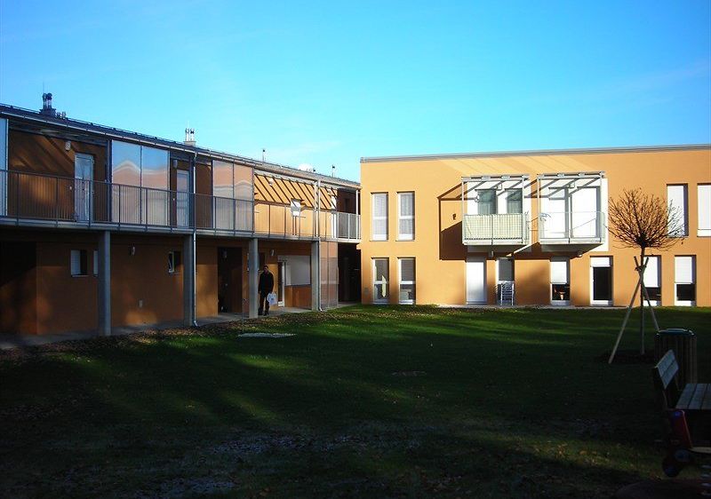 Ein gelbes Apartmentgebäude mit Balkonen und einer Person, die davor geht. Ein grüner Rasen und ein kahler Baum sind ebenfalls sichtbar.