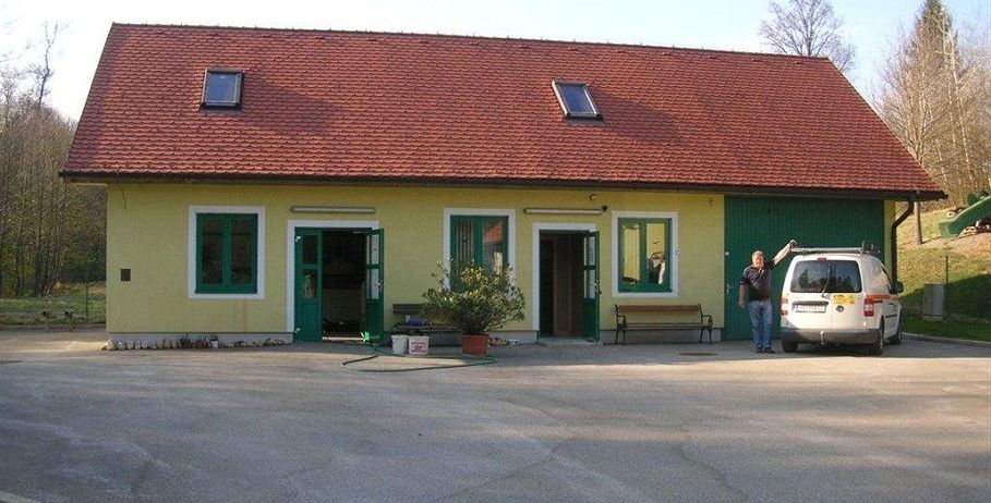 Ein Mann steht neben einem geparkten weißen Van vor einem gelben Haus mit rotem Dach und grüner Umrandung. Das Haus hat mehrere Fenster und zwei offene Türen.