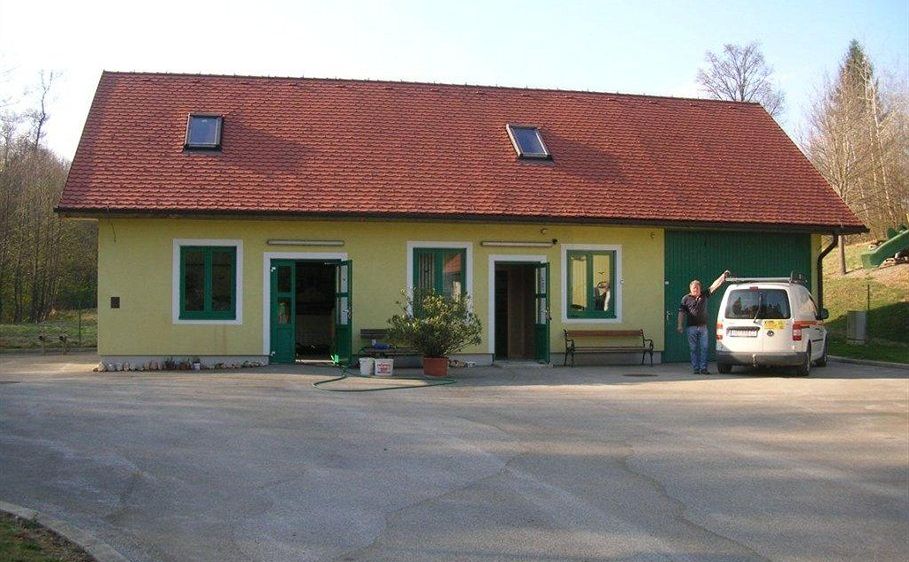 Ein Mann steht neben einem geparkten weißen Van vor einem gelben Haus mit rotem Dach und grüner Umrandung. Das Haus hat mehrere Fenster und zwei offene Türen.