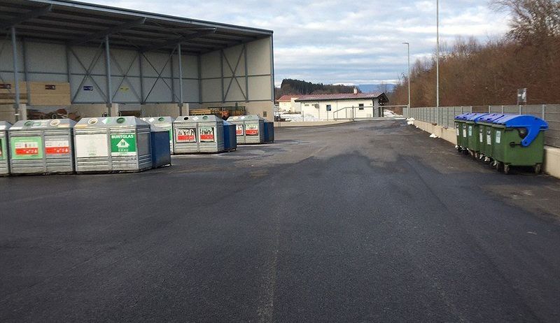 Ein leerer Parkplatz mit mehreren Recyclingbehältern in einer Reihe und einem großen Lagerhaus im Hintergrund.