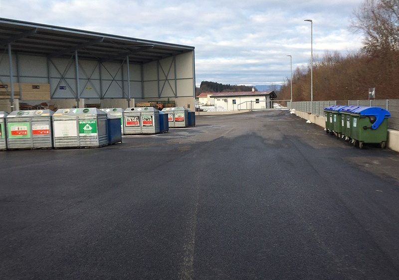Ein leerer Parkplatz mit mehreren Recyclingbehältern in einer Reihe und einem großen Lagerhaus im Hintergrund.