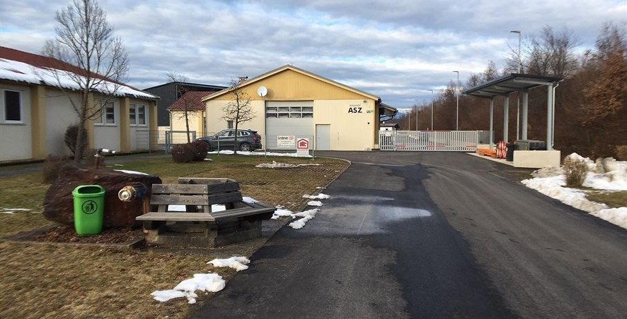 Ein Außenbereich mit einem gelben Gebäude mit der Aufschrift ASZ. Ein Auto ist vor dem Gebäude geparkt. Eine Holzbank steht am Straßenrand. Ein Mülleimer steht am Straßenrand. Rechts vom Weg steht eine Überdachung. Es gibt einige Bäume und Büsche in der Umgebung.