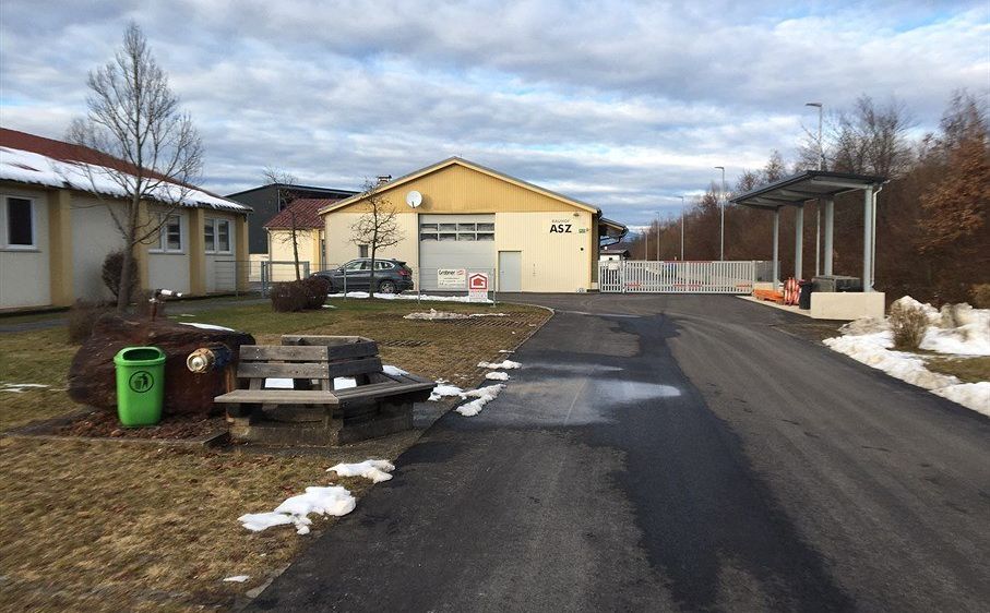 Ein Außenbereich mit einem gelben Gebäude mit der Aufschrift ASZ. Ein Auto ist vor dem Gebäude geparkt. Eine Holzbank steht am Straßenrand. Ein Mülleimer steht am Straßenrand. Rechts vom Weg steht eine Überdachung. Es gibt einige Bäume und Büsche in der Umgebung.