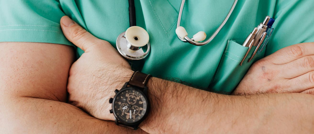 Ein medizinischer Fachmann mit Uhr, Stethoskop und Stift in seiner grünen Arbeitskleidung steht mit verschränkten Armen.