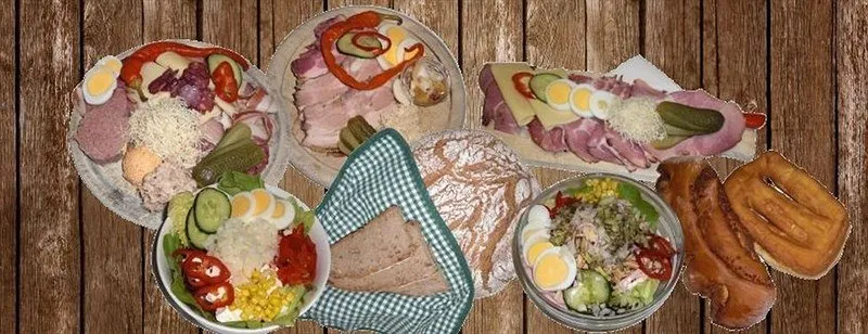 Auf einem Holztisch sind verschiedene Speisen zu sehen. Es gibt Platten mit geschnittenem Fleisch, Eiern, Gurken und Gemüse. Schüsseln enthalten Salate und Mais. Brötchen und Brezeln sind ebenfalls vorhanden.