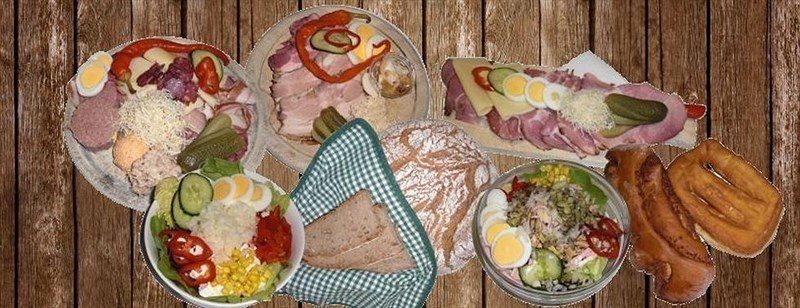 Auf einem Holztisch sind verschiedene Speisen zu sehen. Es gibt Platten mit geschnittenem Fleisch, Eiern, Gurken und Gemüse. Schüsseln enthalten Salate und Mais. Brötchen und Brezeln sind ebenfalls vorhanden.