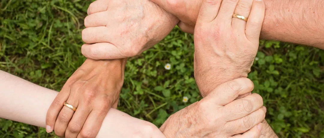 Eine Gruppe von Menschen, sowohl Männer als auch Frauen, halten sich auf einem Grasfeld in einer Handkreishaltung. Sie tragen Eheringe.