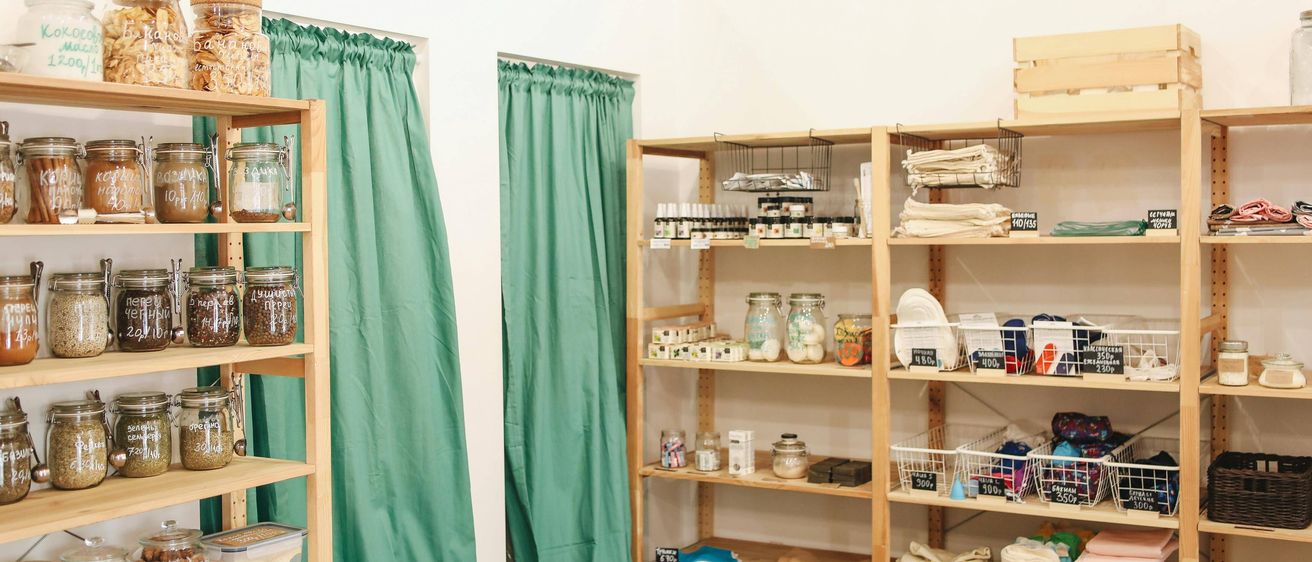 Ein Geschäft mit Holzregalen und türkisen Vorhängen. Die Regale halten Gläser und Behälter mit Etiketten und Preisen. Der Hintergrund ist weiß.