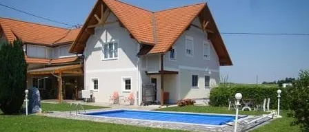 Ein weißes Haus mit rotem Dach und einem Swimmingpool im Garten. Es gibt Pflanzen, Stühle und Lampen auf dem Rasen. Der Himmel ist blau.