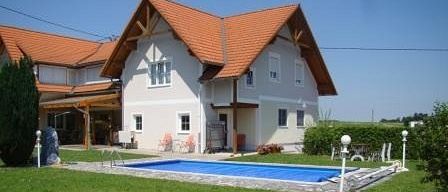 Ein weißes Haus mit rotem Dach und einem Swimmingpool im Garten. Es gibt Pflanzen, Stühle und Lampen auf dem Rasen. Der Himmel ist blau.