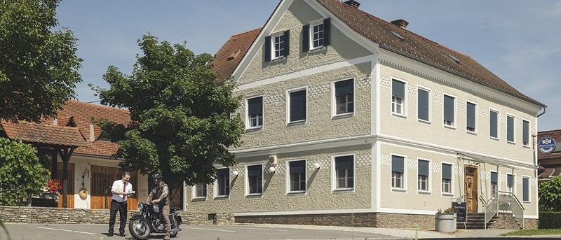 Zwei Personen stehen vor einem Gebäude mit einem Motorrad, das Gebäude hat ein braunes Dach und weiße Wände mit vielen Fenstern.