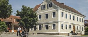 Zwei Personen stehen vor einem Gebäude mit einem Motorrad, das Gebäude hat ein braunes Dach und weiße Wände mit vielen Fenstern.