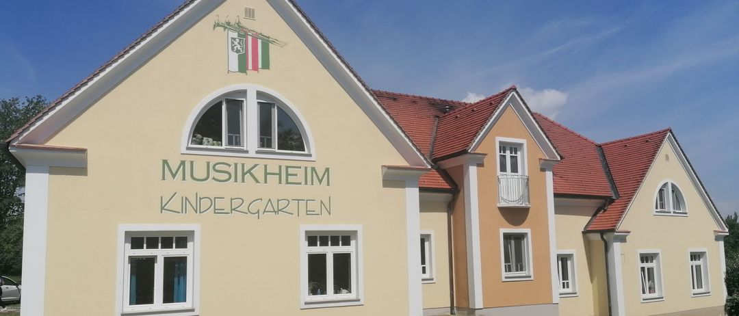 Die Vorderseite eines gelben Gebäudes mit 'MUSIKHEUM KINDERGARTEN' an der Wand, mit einem roten Dach und mehreren Fenstern, unter einem blauen Himmel.