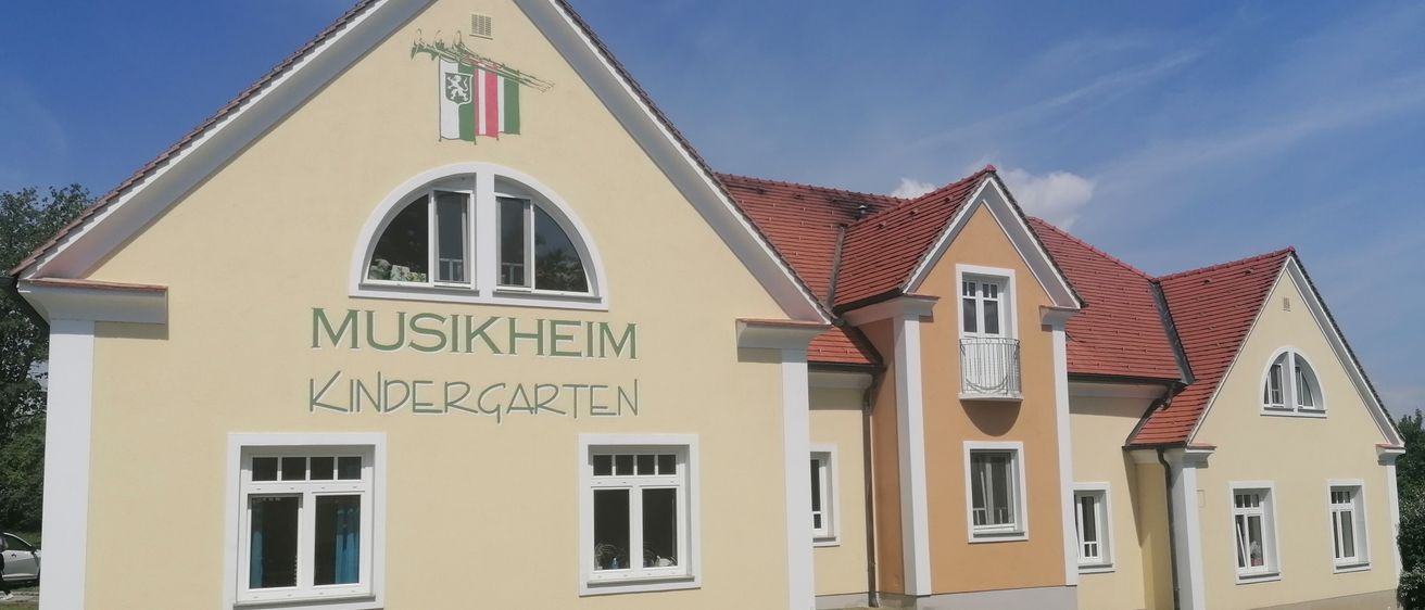 Die Vorderseite eines gelben Gebäudes mit 'MUSIKHEUM KINDERGARTEN' an der Wand, mit einem roten Dach und mehreren Fenstern, unter einem blauen Himmel.