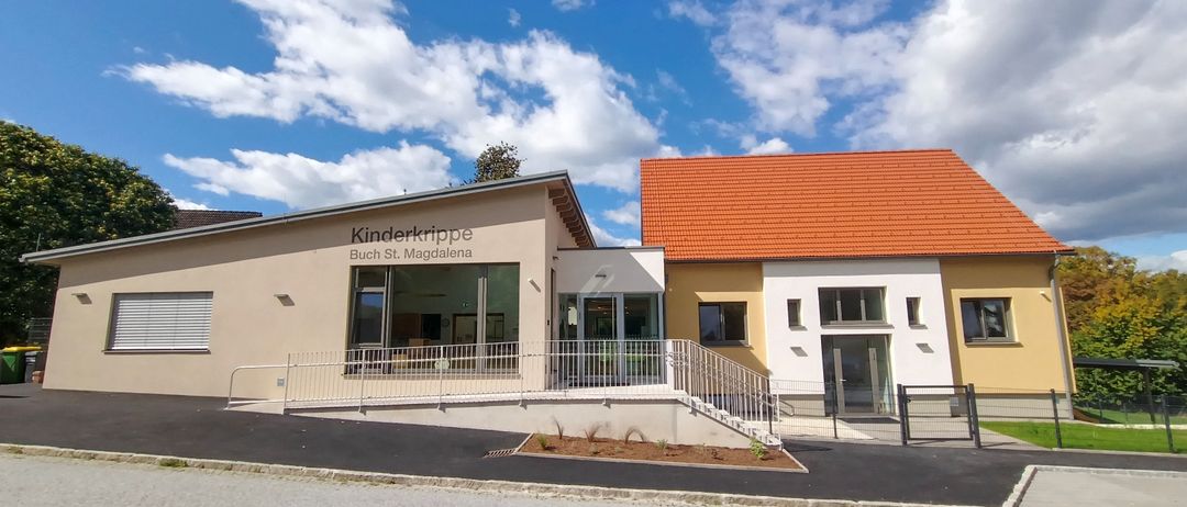 Die Vorderseite eines Gebäudes mit zwei Abschnitten. Links ein Schild mit der Aufschrift 'Kinderkrippe Buch St. Magdalene'. Rechts ein rotes Dach. Oben ein blauer Himmel mit Wolken.