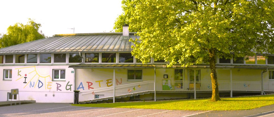 Ein Kindergartengebäude mit einem Metalldach und einem großen Baum davor. Die Wand ist mit dem Wort 'Kindergarten' in bunten Buchstaben bemalt. Es gibt eine Treppe, die zum Eingang führt.