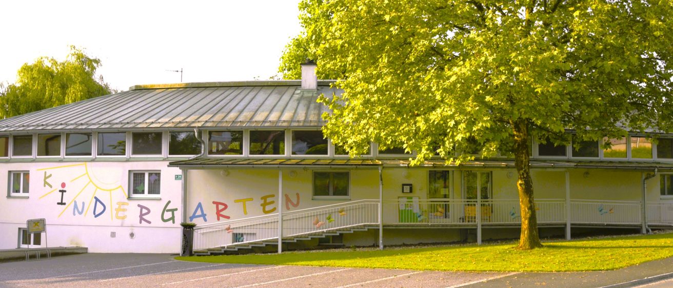 Ein Kindergartengebäude mit einem Metalldach und einem großen Baum davor. Die Wand ist mit dem Wort 'Kindergarten' in bunten Buchstaben bemalt. Es gibt eine Treppe, die zum Eingang führt.