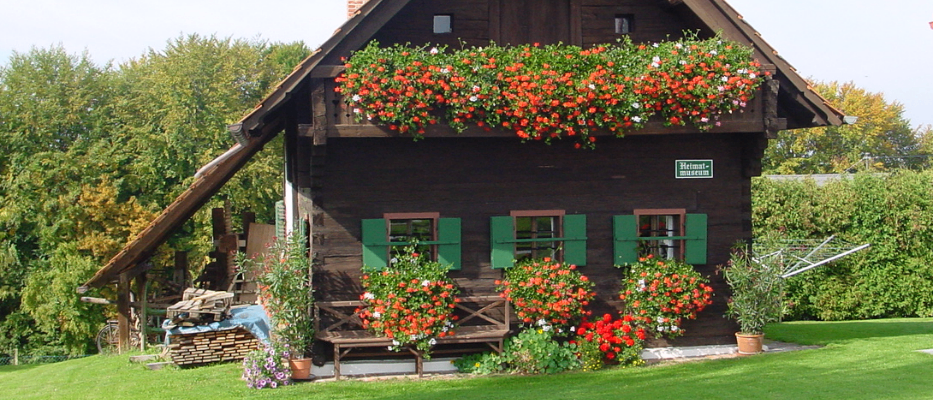 Ein kleines, rustikales Haus mit einem dreieckigen Dach und Ziegelkaminen. Es hat grüne Fensterläden und ist von roten und weißen Blumen umgeben. Vor dem Haus steht eine Holzbank und es gibt Topfpflanzen.