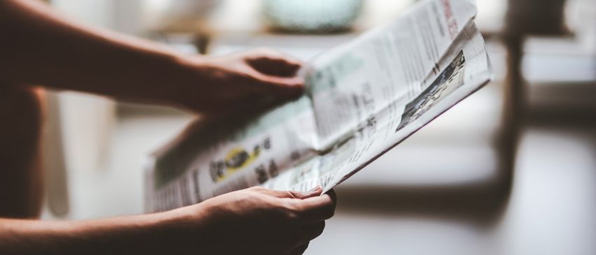 Eine Person hält eine Zeitung in den Händen, möglicherweise liest sie sie. Die Zeitung hat etwas Text und Bilder. Der Hintergrund ist unscharf.