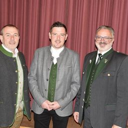 Drei Männer stehen zusammen vor einem roten Vorhang, gekleidet in traditioneller Kleidung. Der Mann auf der linken Seite trägt eine grün-graue Jacke. Der Mann in der Mitte hat eine graue Jacke mit einem weißen Hemd darunter. Der Mann auf der rechten Seite trägt eine grüne Weste über einer grauen Jacke und hat eine Brille.