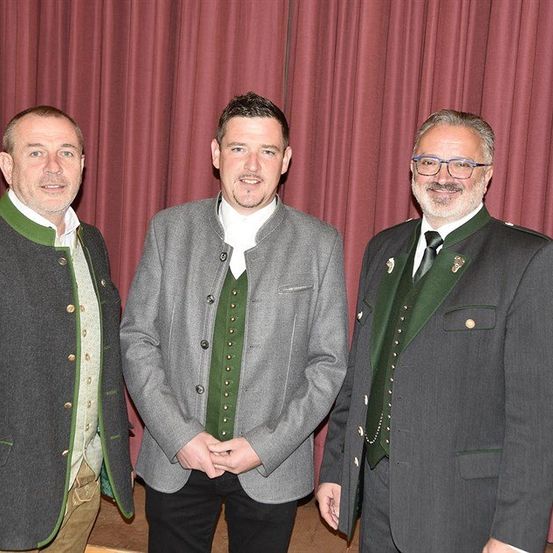 Drei Männer stehen zusammen vor einem roten Vorhang, gekleidet in traditioneller Kleidung. Der Mann auf der linken Seite trägt eine grün-graue Jacke. Der Mann in der Mitte hat eine graue Jacke mit einem weißen Hemd darunter. Der Mann auf der rechten Seite trägt eine grüne Weste über einer grauen Jacke und hat eine Brille.