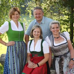 Vier Menschen stehen in einem Park, gekleidet in traditionelle deutsche Kleidung. Zwei Frauen und ein Mann lächeln. Der Mann steht hinter den Frauen.