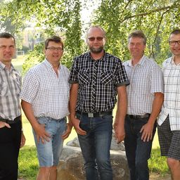 Fünf Männer stehen in einer Reihe unter einem Baum, lächeln und schauen in die Kamera. Sie sind in Freizeitkleidung gekleidet, und einige tragen eine Brille. Hinter ihnen sind Bäume, ein großer Felsbrocken und ein Grasbereich. In der Ferne befindet sich ein Gebäude und ein Zaun.