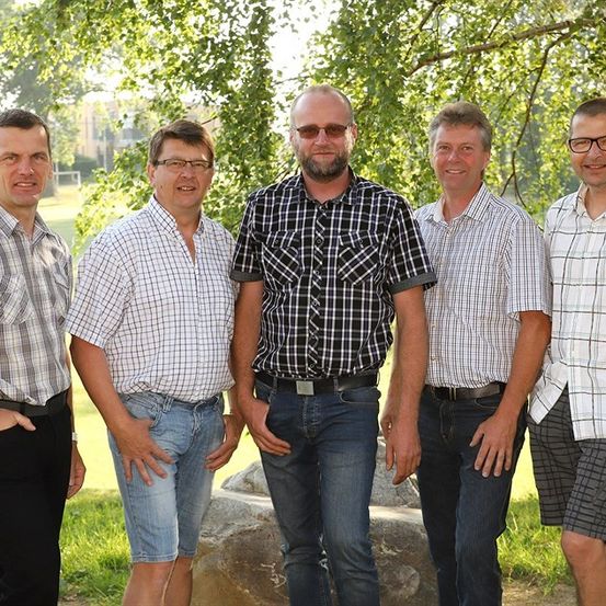 Fünf Männer stehen in einer Reihe unter einem Baum, lächeln und schauen in die Kamera. Sie sind in Freizeitkleidung gekleidet, und einige tragen eine Brille. Hinter ihnen sind Bäume, ein großer Felsbrocken und ein Grasbereich. In der Ferne befindet sich ein Gebäude und ein Zaun.