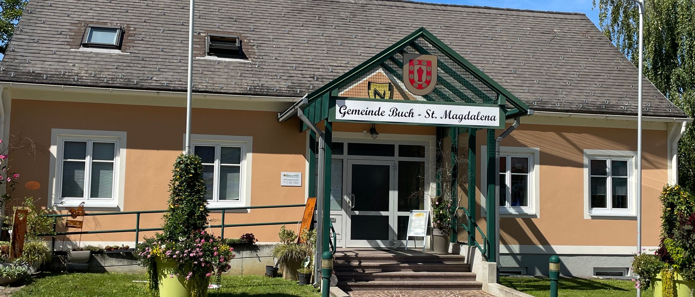 Ein Gebäude mit grünem Dach und Eingang, beschriftet mit Gemeinde Buch - St. Magdalena, hat mehrere Fenster, einen Fahnenmast und ist von Pflanzen und Gras umgeben.