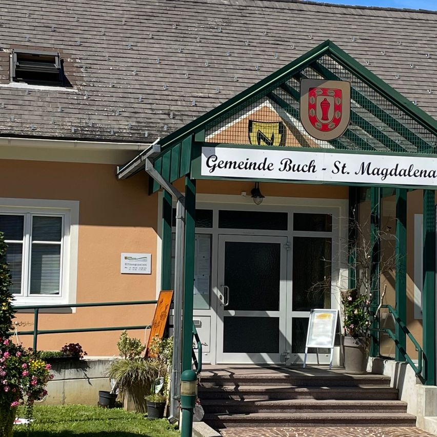 Ein Gebäude mit grünem Dach und Eingang, beschriftet mit Gemeinde Buch - St. Magdalena, hat mehrere Fenster, einen Fahnenmast und ist von Pflanzen und Gras umgeben.