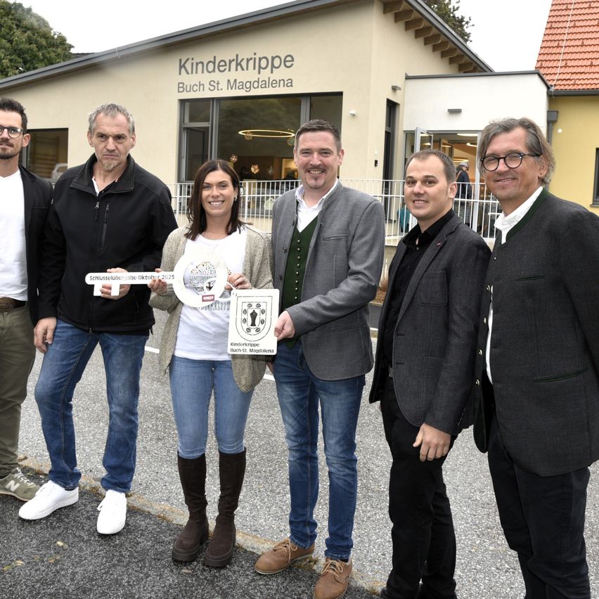 Fünf Personen stehen vor einem Gebäude. Die Frau hält ein Zertifikat und einen Schlüssel. Der Schriftzug auf dem Gebäude lautet 'Kinderkrippe Buch St. Magdalena'.