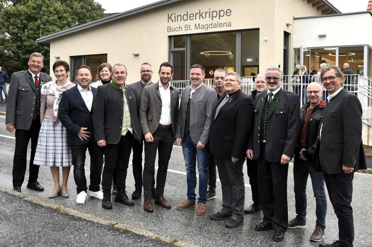 Eine Gruppe von Männern und Frauen steht vor einem Gebäude mit dem Schild 'Kinderkrippe Buch St. Magdalena.' Sie sind in formeller Kleidung gekleidet, einige mit Brillen und andere mit Lächeln.