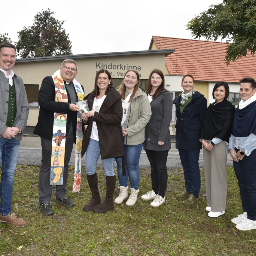 Eine Gruppe von Menschen steht vor dem Kinderkrippe-Gebäude. Eine Person hält ein Buch und lächelt. Sie sind in Freizeitkleidung gekleidet und stehen auf Gras.