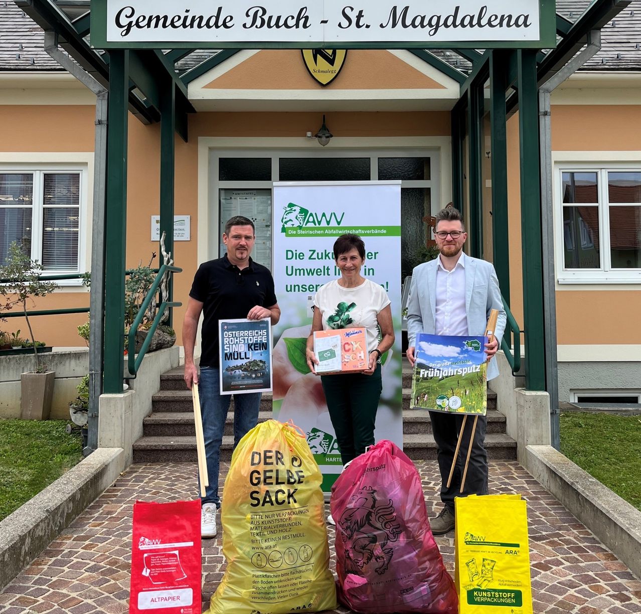 Drei Personen stehen vor einem Gebäude mit einem Schild, das Gemeinde Buch-St. Magdalena liest. Sie halten Bücher und gelbe Taschen. Der Mann links hat ein Poster über Recycling, die Frau in der Mitte hat ein Buch und eine Tasche, und der Mann rechts hat ein Poster und eine Tasche.